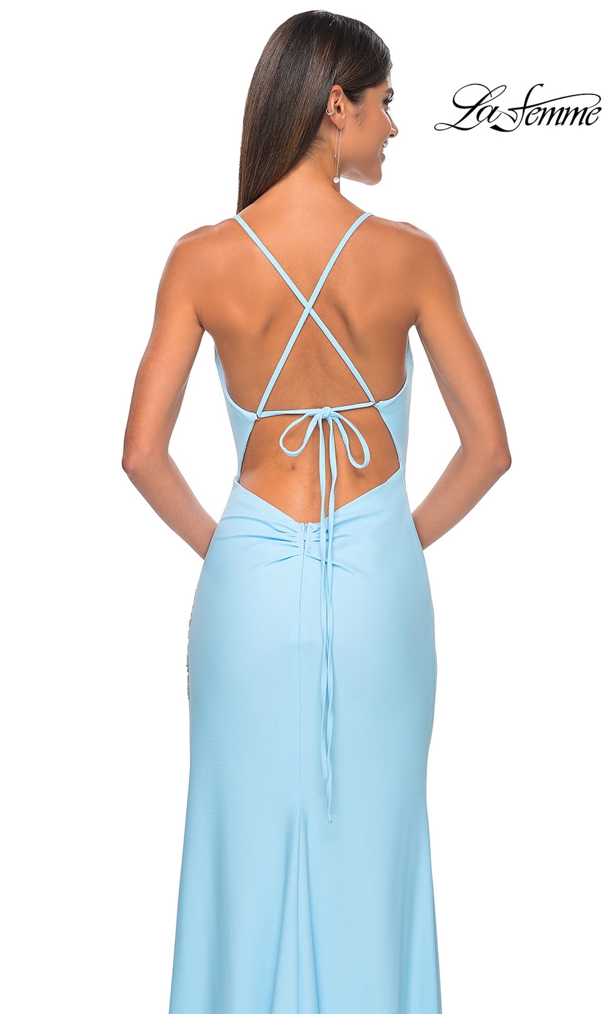 A woman wearing the La Femme Cowl-Neck Pastel Jersey Prom Dress 31574, a light blue floor-length gown with cowl neckline, low back, crisscross straps, and tie detail, stands against a white background.